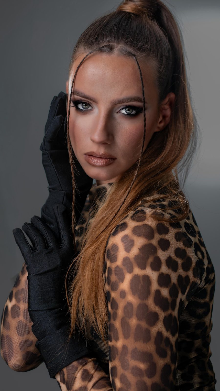 Model met strakke hoge staart, zwarte handschoenen en luipaardprint, warme bronzen ogen, voor een grijze fotoachtergrond